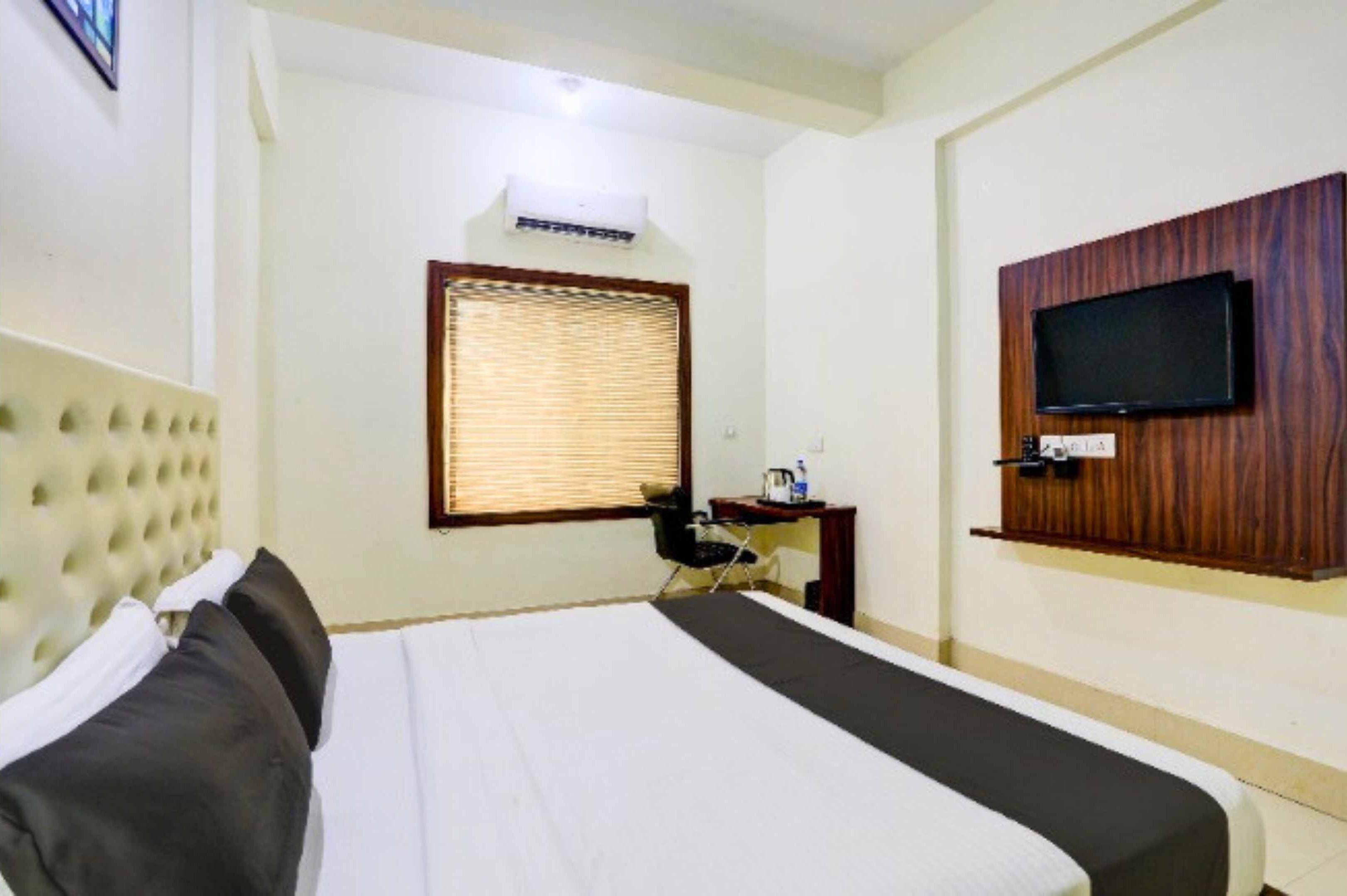 Ein Hotelzimmer mit einem großen Bett, einem Schreibtisch, einem an der Wand befestigten Fernseher und einem Fenster mit Jalousien.