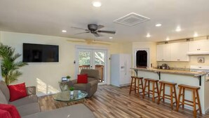 A living room with a sofa, a glass coffee table, and a ceiling fan. There is a kitchen with a refrigerator, a window, and wooden stools.