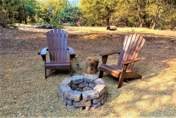Two Adirondack chairs and a stone fire pit in a forested area.