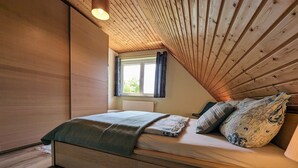 A bedroom with a wooden ceiling, a bed with patterned bedding, a window with curtains, and a radiator. - Wohnung auf Usedom mit Seeblick (Usedom)