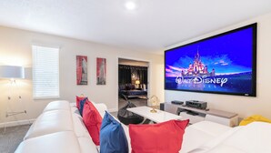 Een moderne woonkamer met een witte bank, rode en blauwe kussens, een flatscreen televisie waarop 'Walt Disney Pictures' te zien is, en een kleine tafel met een lamp.