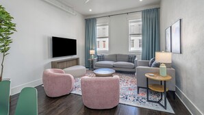 Ein modernes Wohnzimmer mit Flachbildfernseher, einem grauen Sofa, zwei rosafarbenen Sesseln, einem Couchtisch und einem kleinen Beistelltisch mit Lampe.