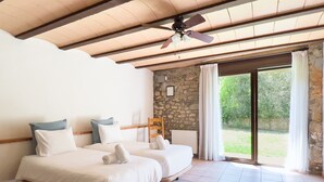 A bedroom with two beds, a stone wall, a ceiling fan, and a view of greenery outside. - Majordoms Molí - La Garrotxa (Beuda)