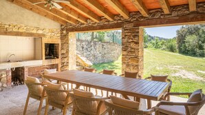An outdoor patio with a stone wall, a wooden table, wicker chairs, and a stone fireplace. - Majordoms Molí - La Garrotxa (Beuda)