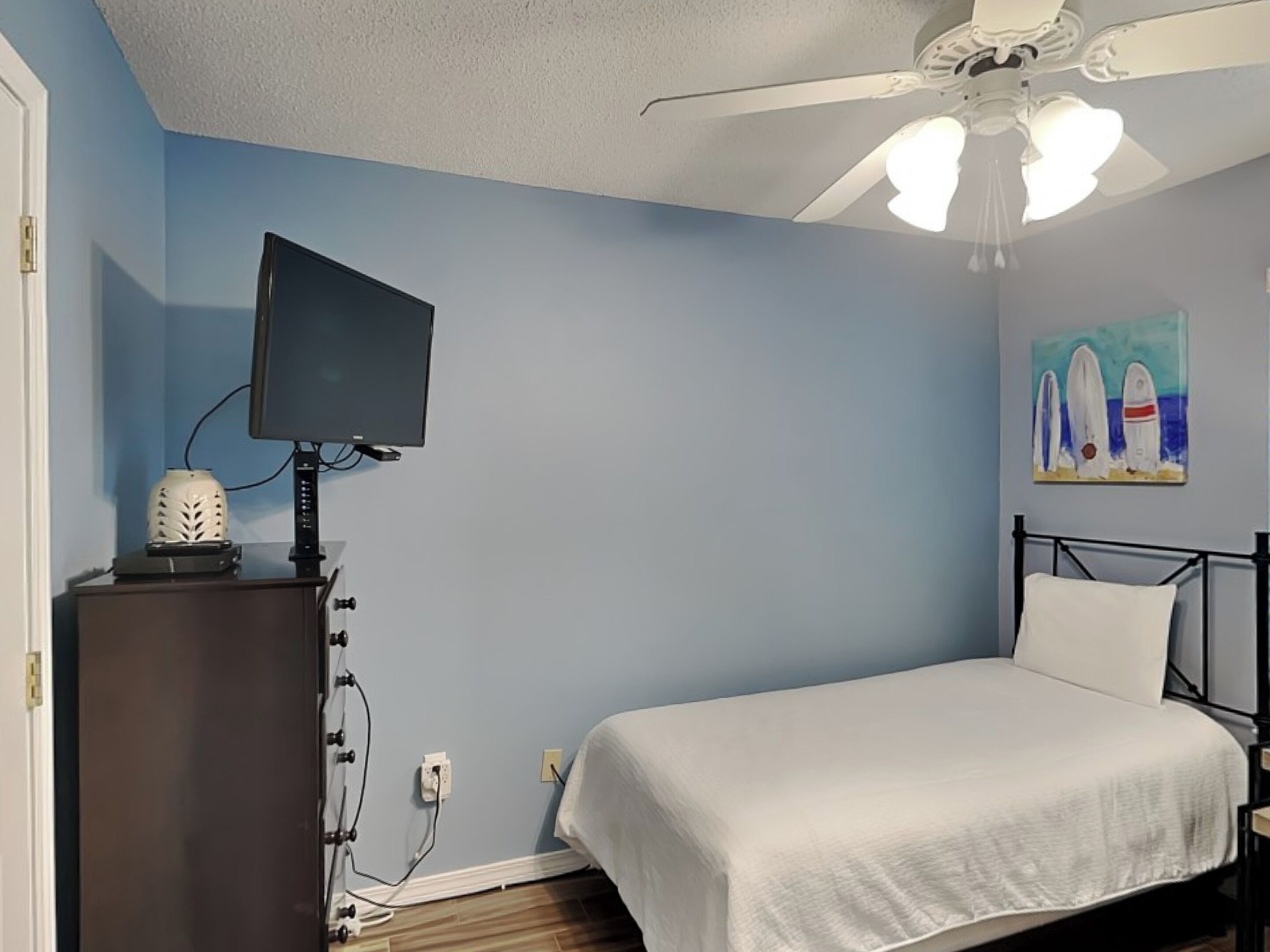 A bedroom with a bed, a ceiling fan, a TV on a stand, and a painting on the wall.