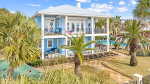 Une maison à deux étages avec une piscine, entourée de palmiers et surplombant un ciel d’un bleu limpide.