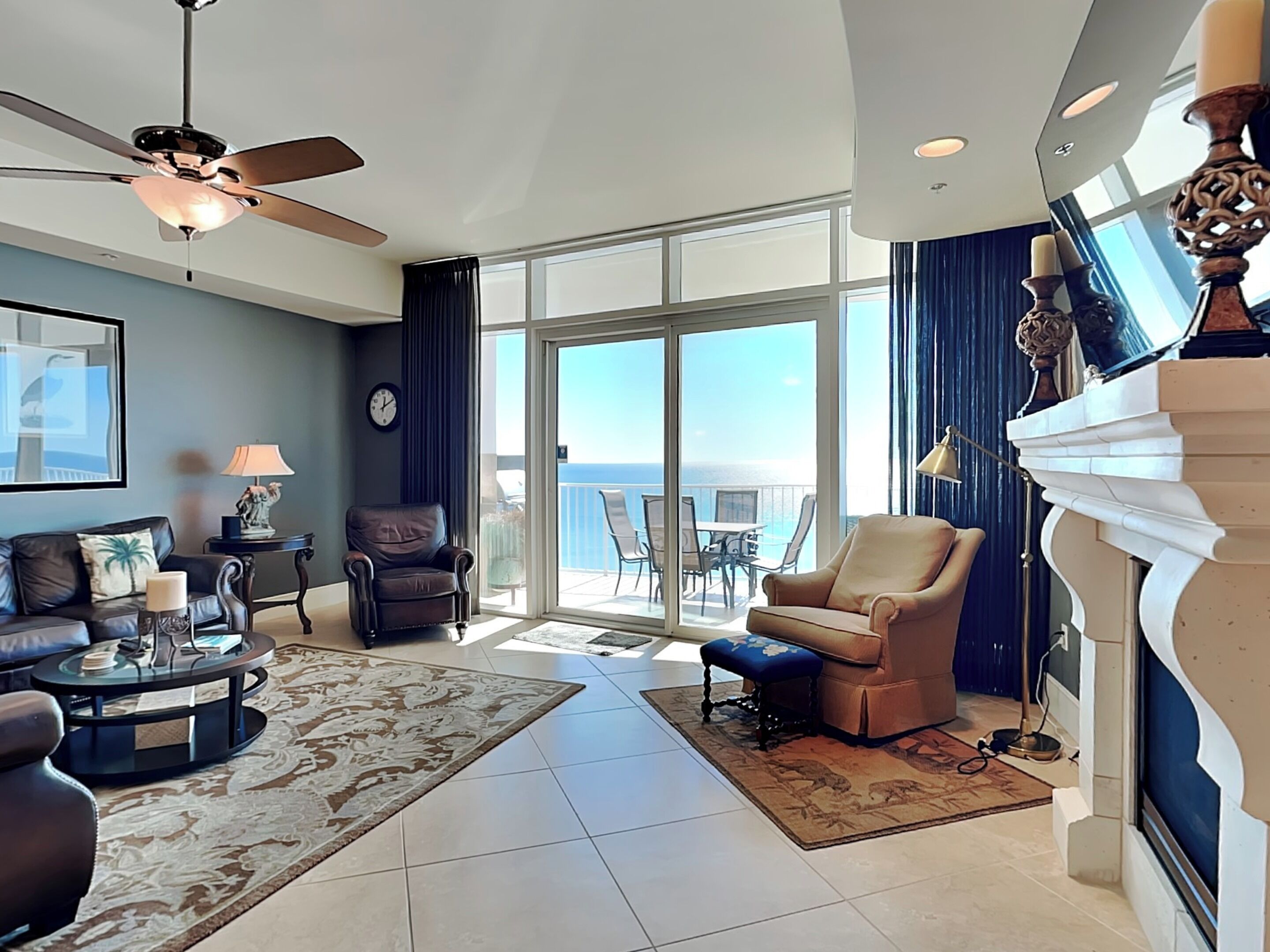 A spacious living room with a large window offering a view of the ocean, a ceiling fan, and a fireplace with a decorative mantel.