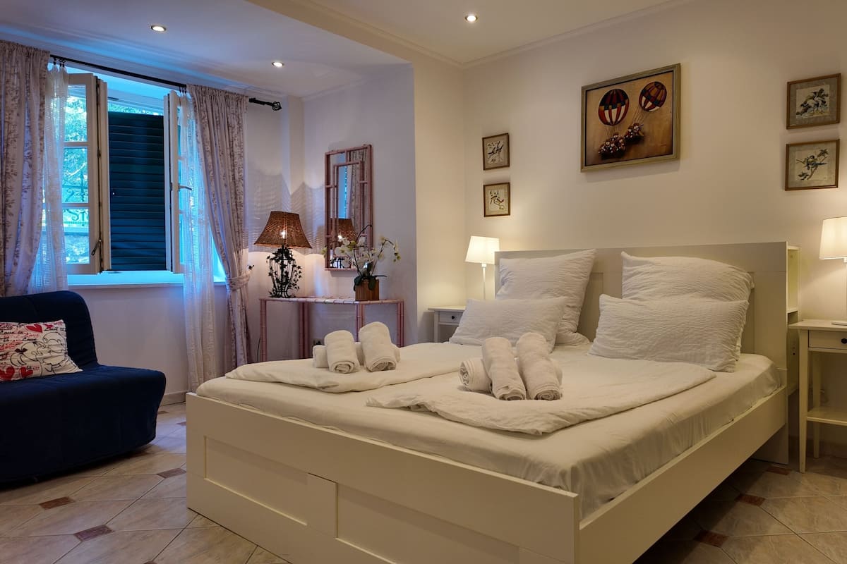 Una cama bien hecha con sábanas y toallas blancas, una mesita de noche con lámpara, una silla con cojín y una ventana con cortinas.