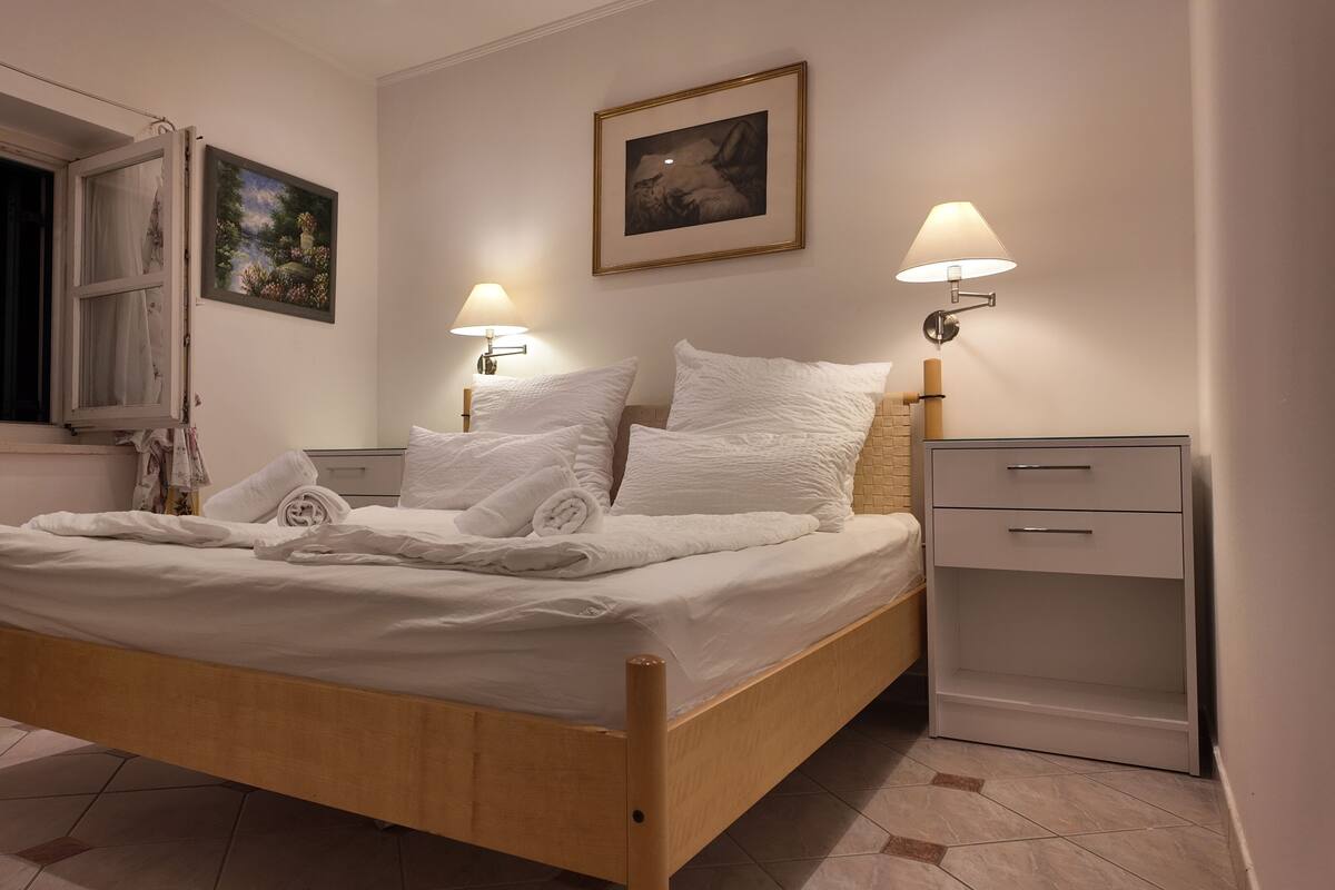 Una cama bien hecha con ropa de cama y toallas blancas, una mesita de noche con una lámpara, un cuadro en la pared y una ventana con cortinas.