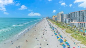A beach with a long stretch of umbrellas and sun loungers, adjacent to a high-rise hotel with multiple floors and balconies. - Sand Dunes 1246 (Myrtle Beach)