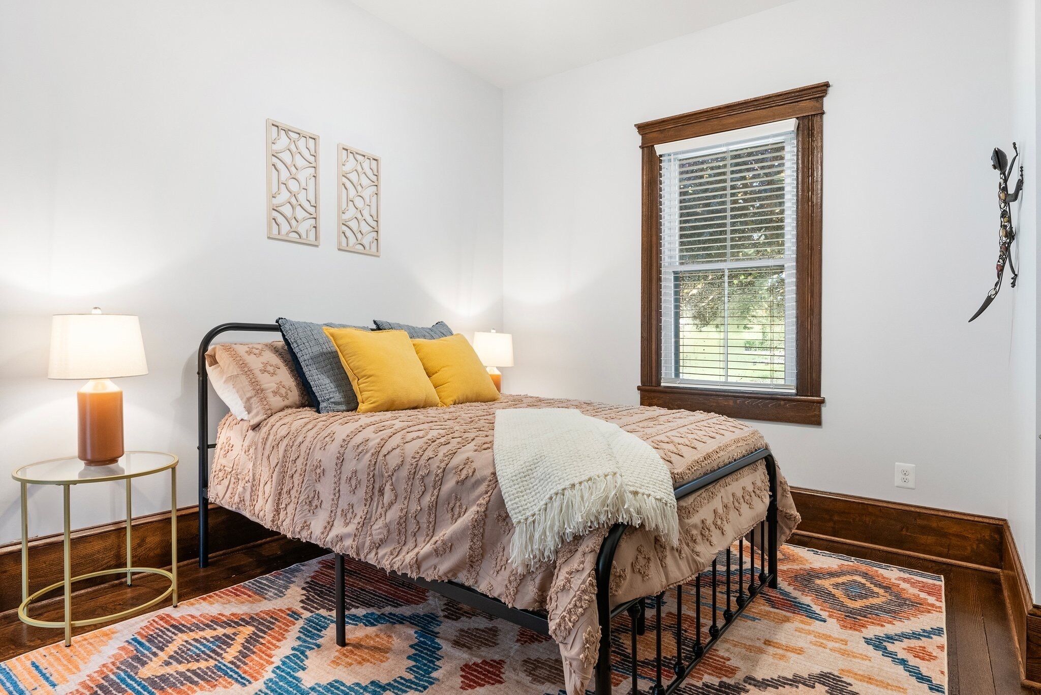 A bedroom with a bed, a window with blinds, a nightstand with a lamp, and decorative wall art.