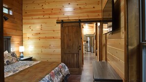 Habitació d'una cabana de fusta amb un llit, una panxa, una tauleta de nit i una porta que dona a una zona de cuina.