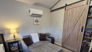 Uma sala de estar acolhedora com um sofá cinzento, uma mesinha com abajur, uma porta de correr estilo celeiro e um ar-condicionado fixado na parede.