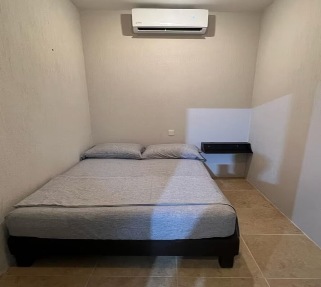 A small, narrow room with a bed, a wall-mounted air conditioner, and a small table with a black surface.