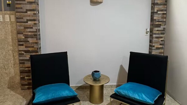 Une pièce comprenant deux chaises noires à l’armature dorée et aux coussins bleus, une petite table ronde sur laquelle se trouve un vase bleu, et une fenêtre donnant sur des arbres.