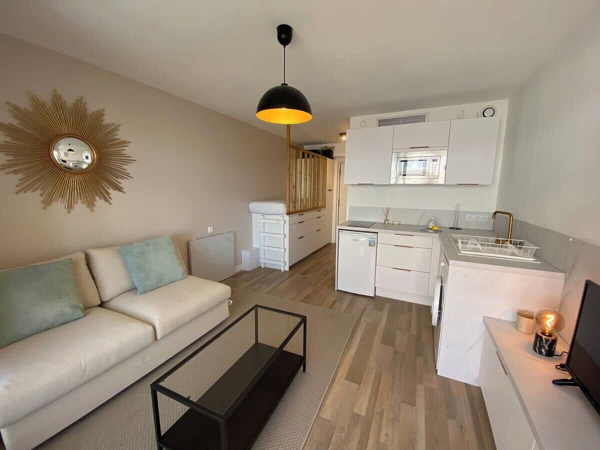 A compact living space with a sofa, coffee table, kitchenette, and a decorative sunburst mirror.