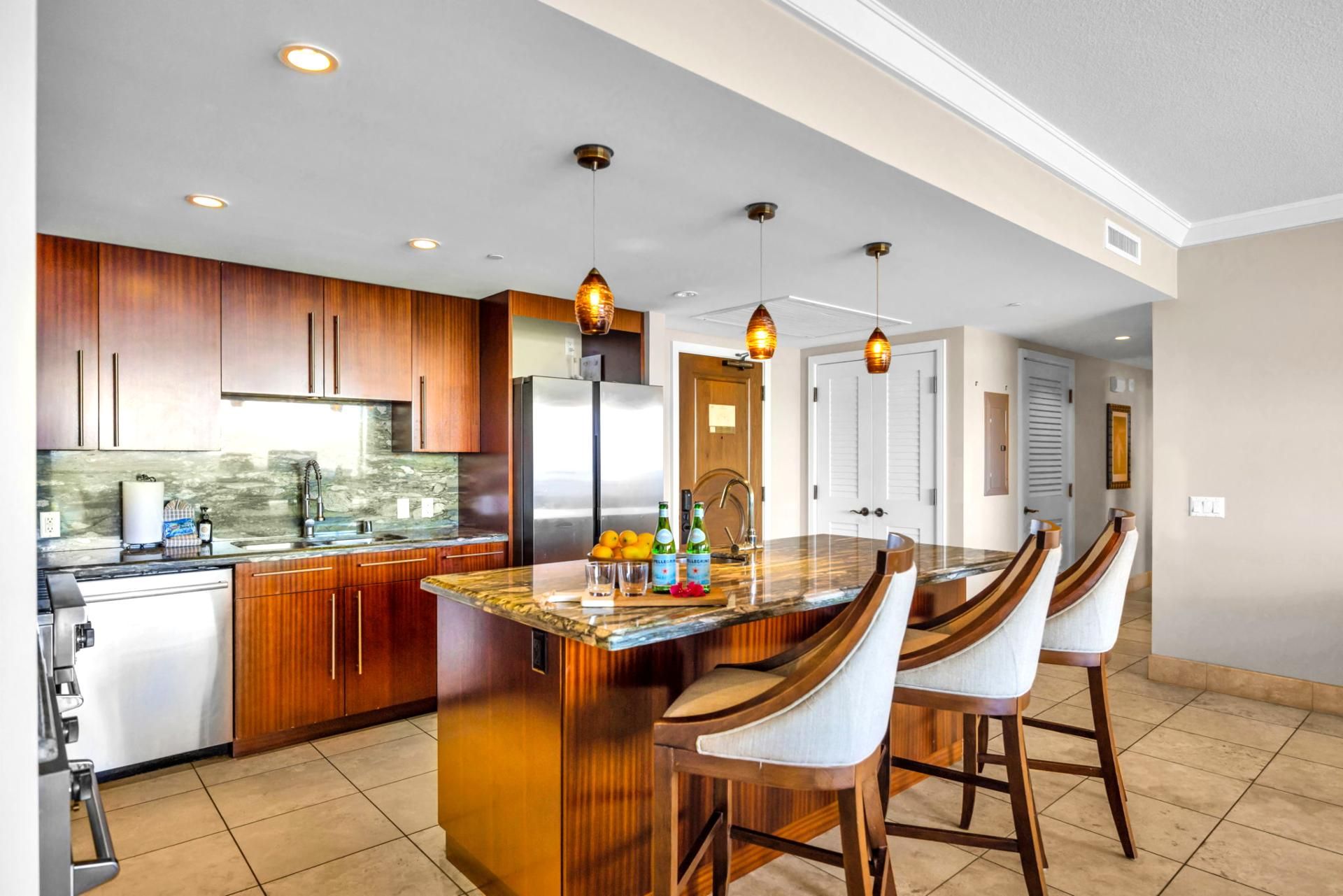 A modern kitchen with wooden cabinets, a stainless steel refrigerator, a marble countertop, and a bar area with bar stools.