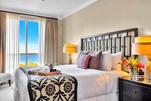 Ein Hotelzimmer mit einem großen Bett, zwei Nachttischlampen, einem Nachttisch mit Blumenvase und Blick auf das Meer durch das Fenster.
