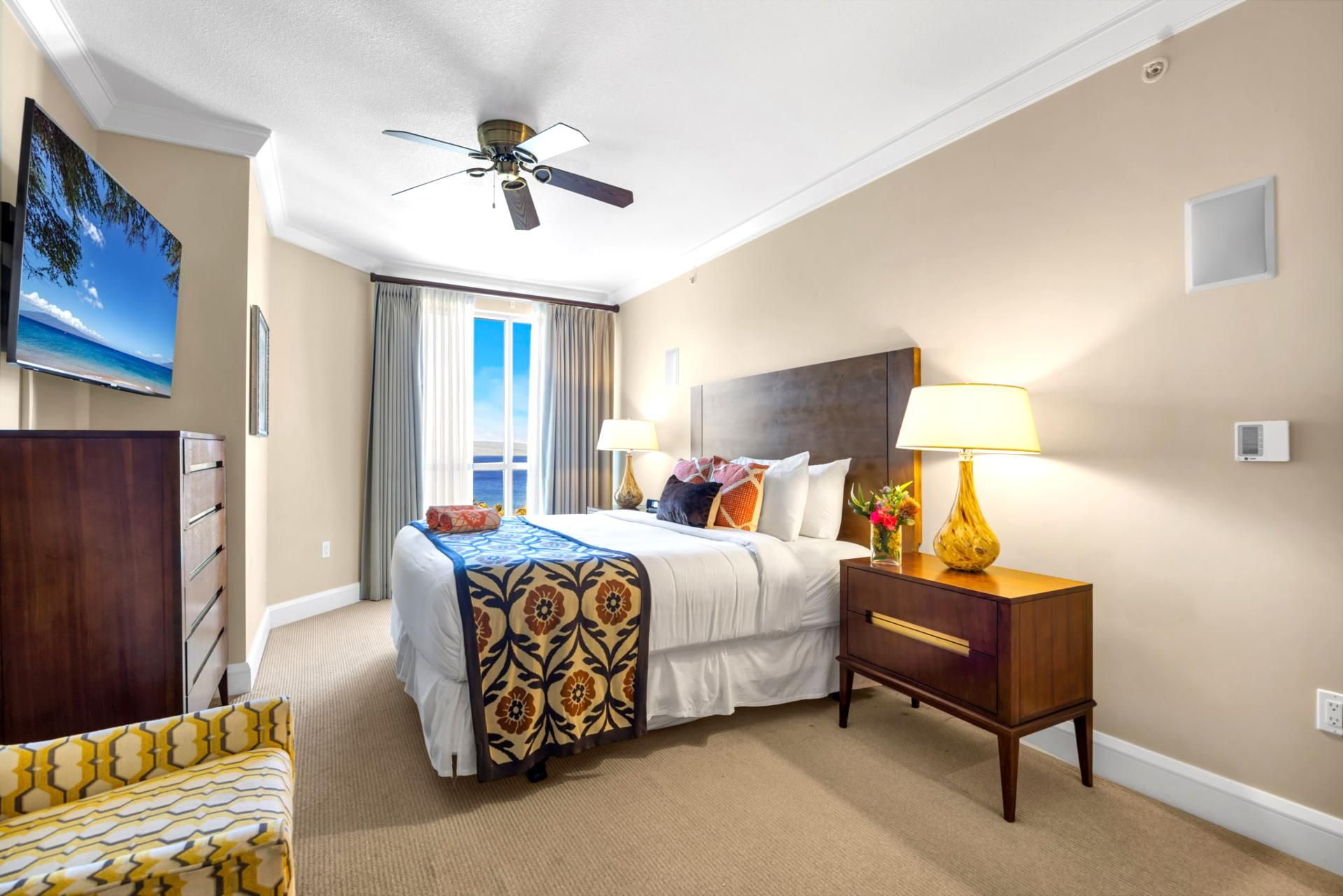A hotel room with a bed, a nightstand with a lamp, a ceiling fan, a TV on the wall, and a window with curtains.