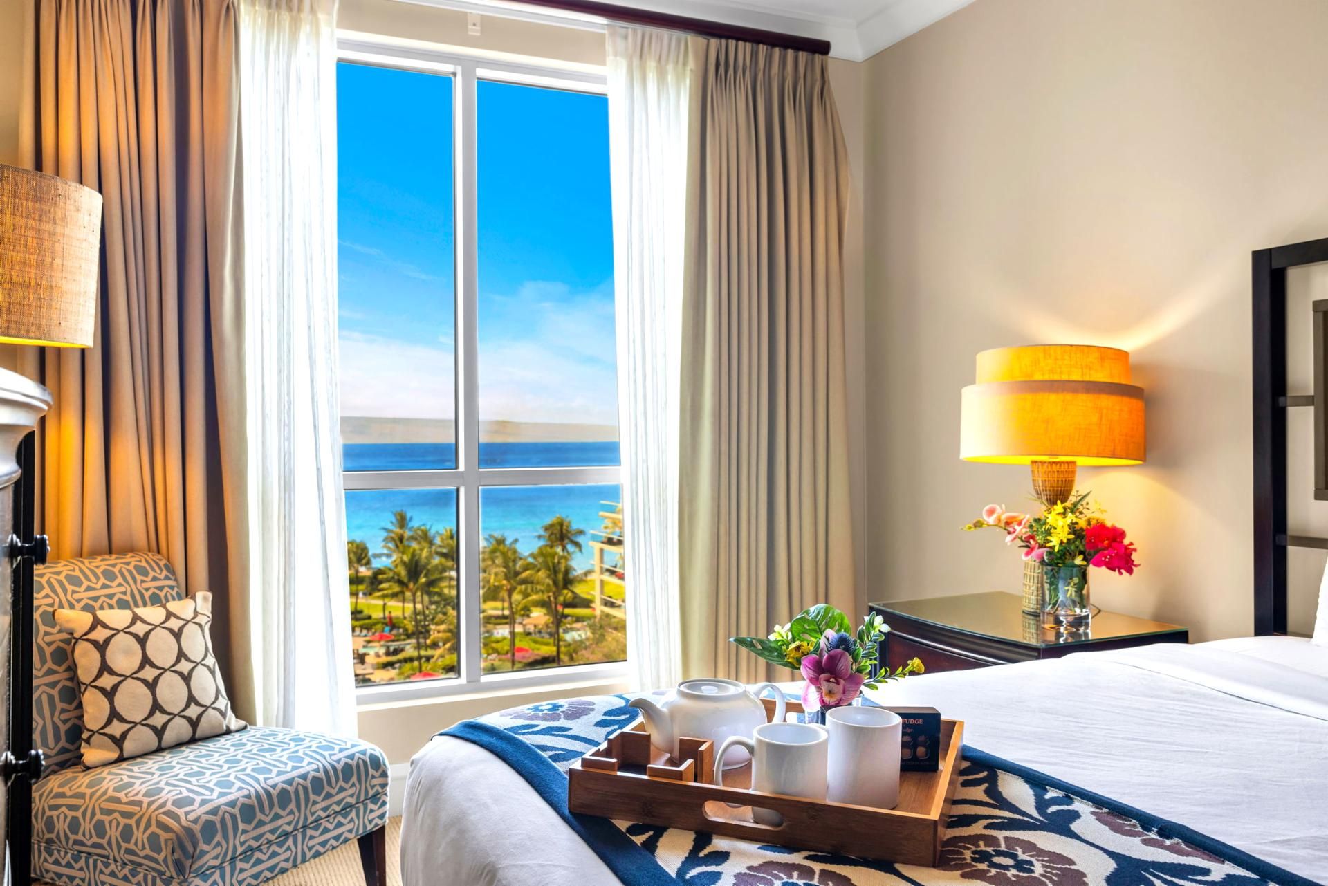 A hotel room with a large window offering a view of the ocean, a bed with a blue and white patterned bedspread, a bedside table with a lamp and a vase of flowers, and a tray with tea set and a remote control.
