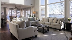 A spacious living room with large windows, a sofa set, a coffee table, and a dining area with a view of the water. - Cimmaron Bay Retreat (Sunrise Beach)