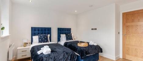 A bedroom with two beds, each with a dark blue headboard and matching bedding. There are bedside tables with lamps, a wooden door, and a small shelf with a plant.