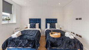 A hotel room with two beds, each with a dark blue headboard and white bedding. There is a window on the left side, and a small basket with items on the right side of the bed. - Flat 1 Saarb Apartments  (Leatherhead)