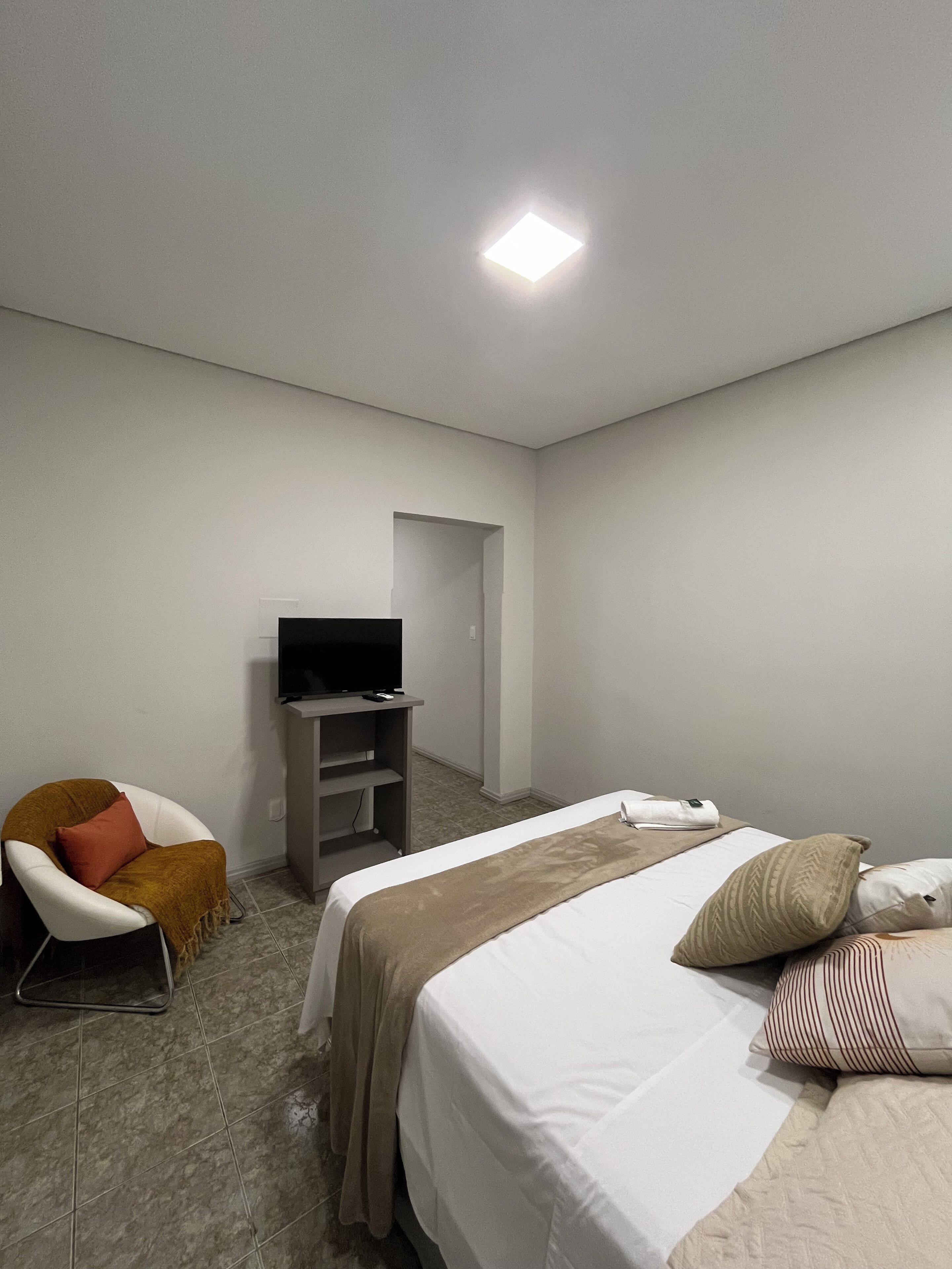 A hotel room with a bed, a TV on a stand, a chair, and a door leading to another room.