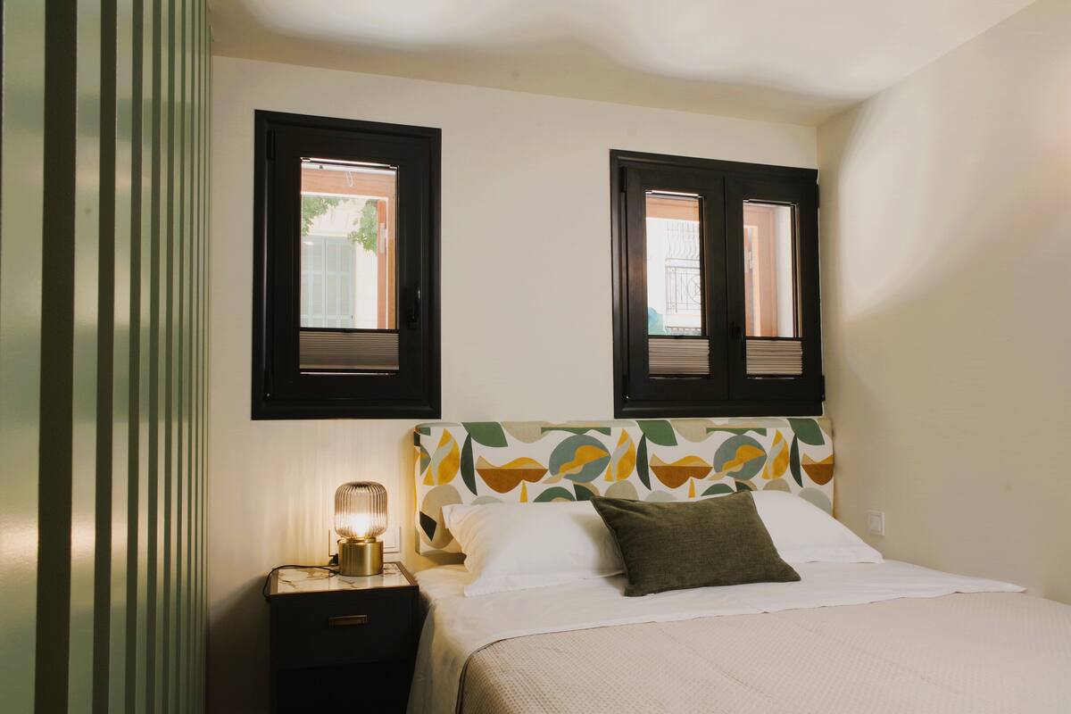 Cama bem arrumada com lençóis brancos e um travesseiro verde, uma luminária na mesa de cabeceira e um criado-mudo com luminária.