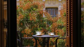 Un balcon avec une table dressée pour deux personnes, entouré d’une végétation luxuriante et, en arrière-plan, un bâtiment agrémenté d’éléments décoratifs.