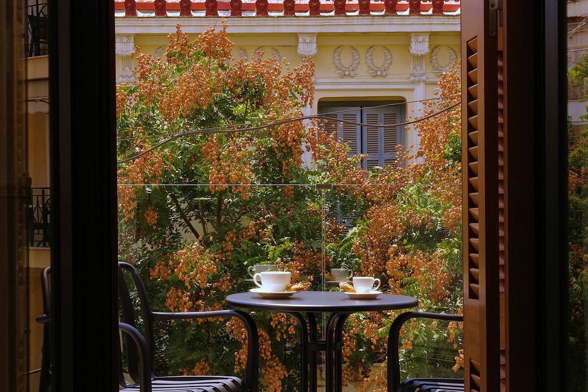 Uma varanda com mesa posta para dois, rodeada por vegetação exuberante e um edifício com detalhes decorativos ao fundo.