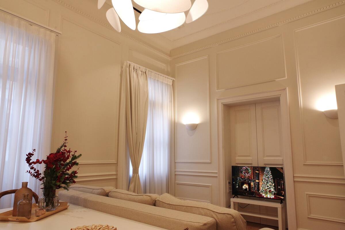 Sala de estar bem iluminada, com um sofá, uma televisão exibindo uma cena natalina e um vaso decorativo com flores sobre uma mesa.