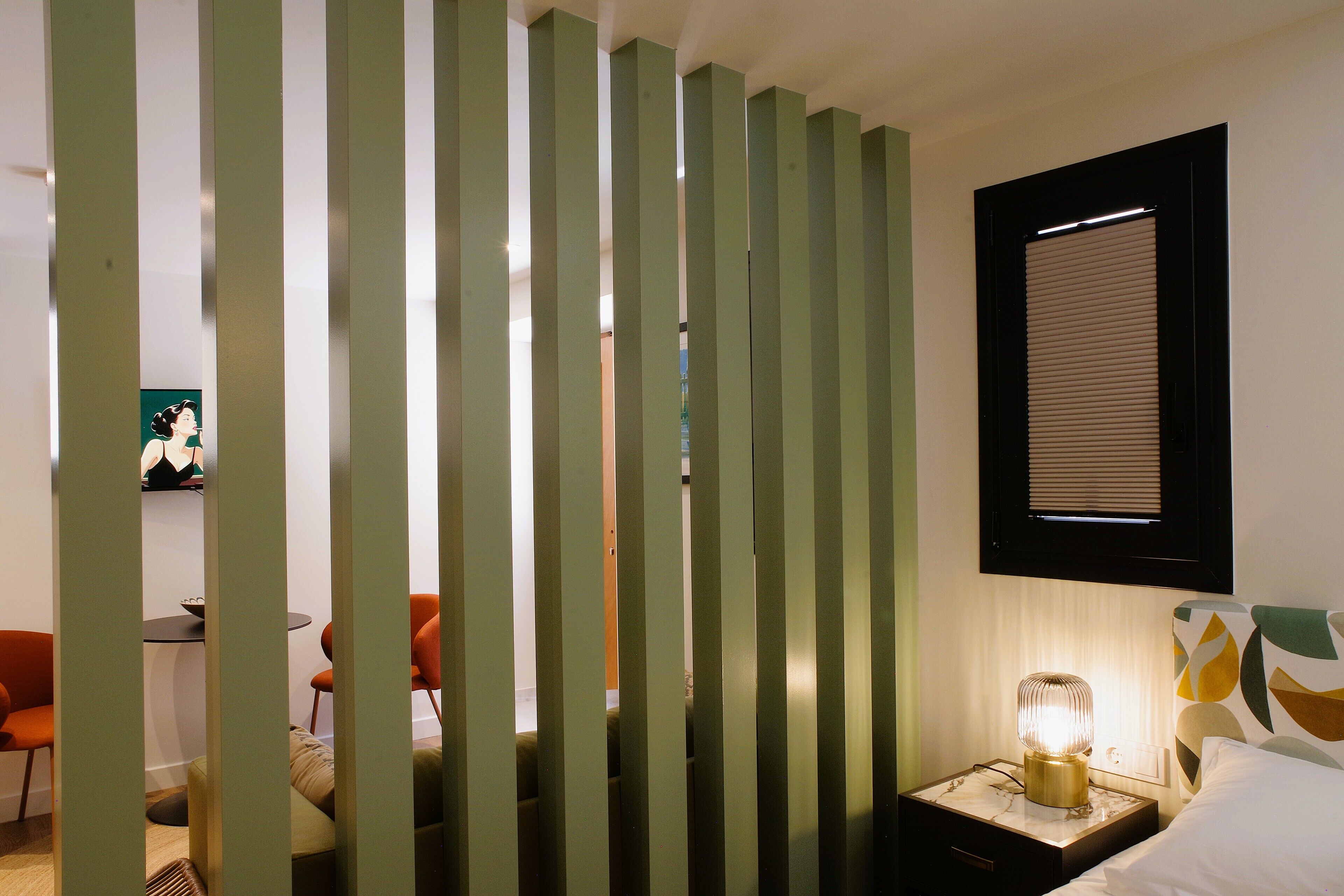Ein Zimmer mit einem Bett, einem Nachttisch mit Lampe, einem Fenster mit Jalousien und einem Sessel.