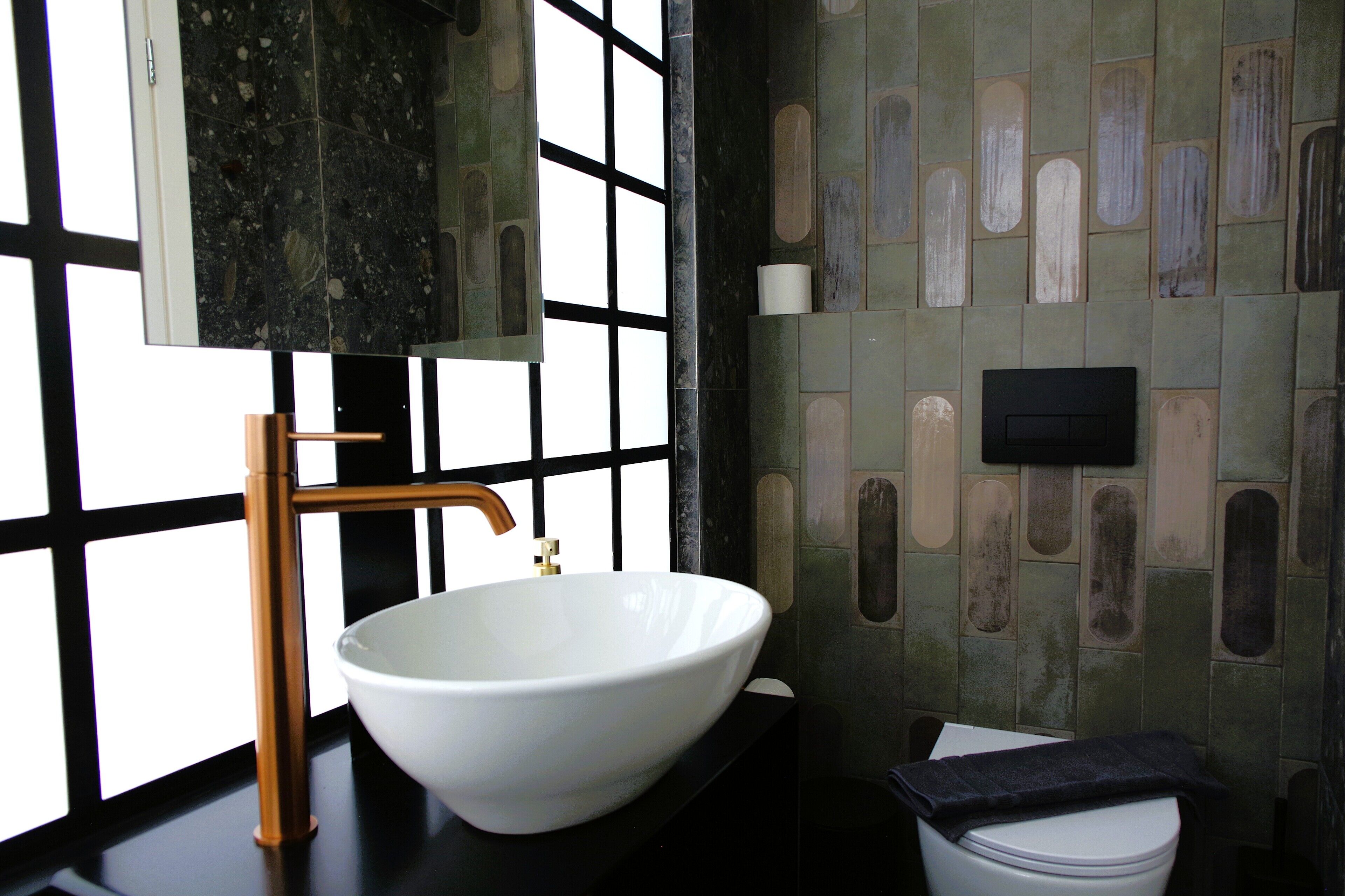 A bathroom with a white vessel sink, a wooden faucet, a black countertop, a toilet, and a tiled wall with rectangular patterns.
