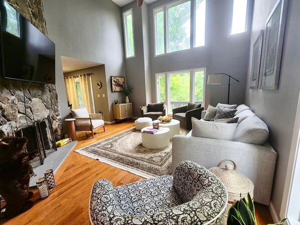 A modern living room with a stone wall, a fireplace, a large TV, and a cozy seating area with a patterned ottoman and a grey sofa.