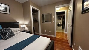 Une chambre d’hôtel avec un lit, une table de chevet, un miroir et un espace pour faire la lessive, visible à travers une porte ouverte.