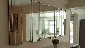 A dining area with a long table, two upholstered chairs, a vase with dried flowers, and a large mirror on the wall.
