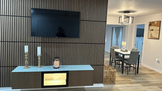 A modern living room with a large flat-screen TV mounted on a wooden panel wall, a glass-top coffee table with candles, and a dining area with a round table and chairs.
