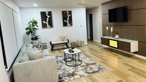 A modern living room with a sofa, armchair, and a coffee table. There is a TV mounted on a wooden wall, a ceiling fan, and a plant.