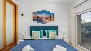 Quarto de hotel com uma cama grande, duas mesas de cabeceira, um quadro de uma praia e vista para o exterior.