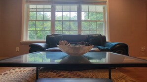 Ein Wohnzimmer mit einem Glastisch, einem dunklen Ledersofa und einem Fenster mit Blick auf Bäume.