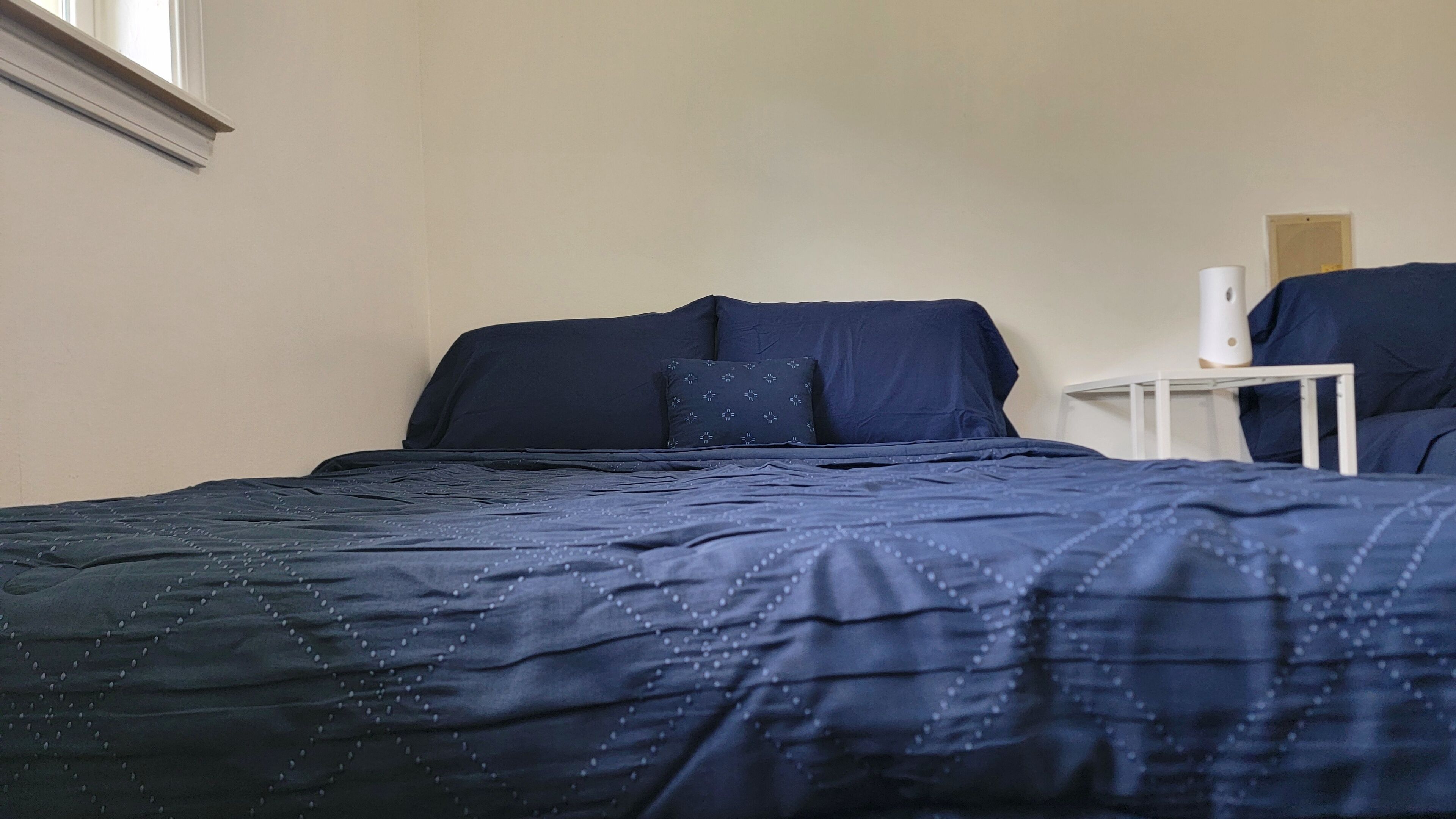 Una cama bien hecha con colcha y almohadas azul oscuro, una mesita de noche con una lámpara blanca, y una ventana con persianas.