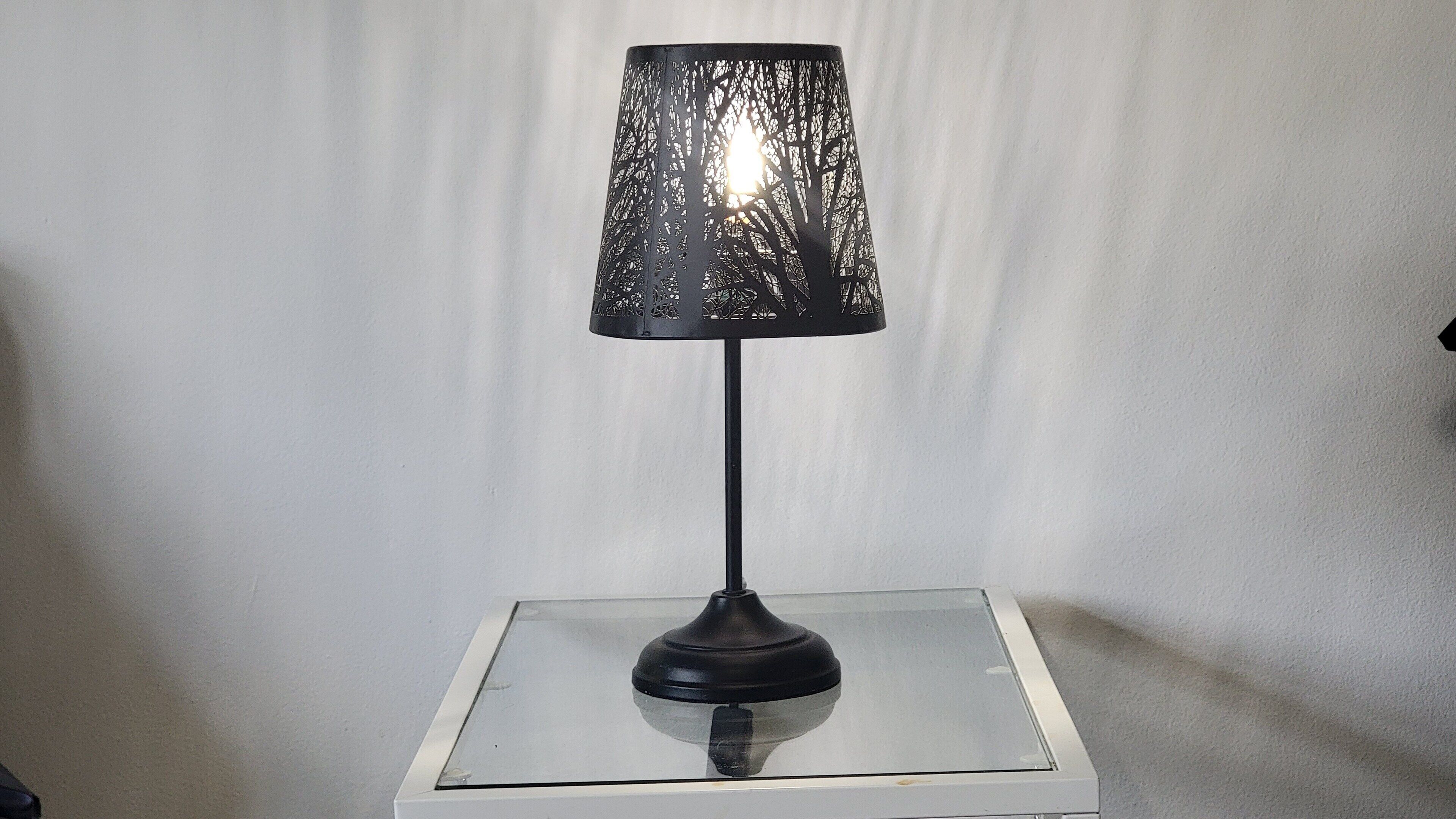 Eine Tischlampe mit schwarter Schirm auf einem Glastisch vor einer schlichten Wand.