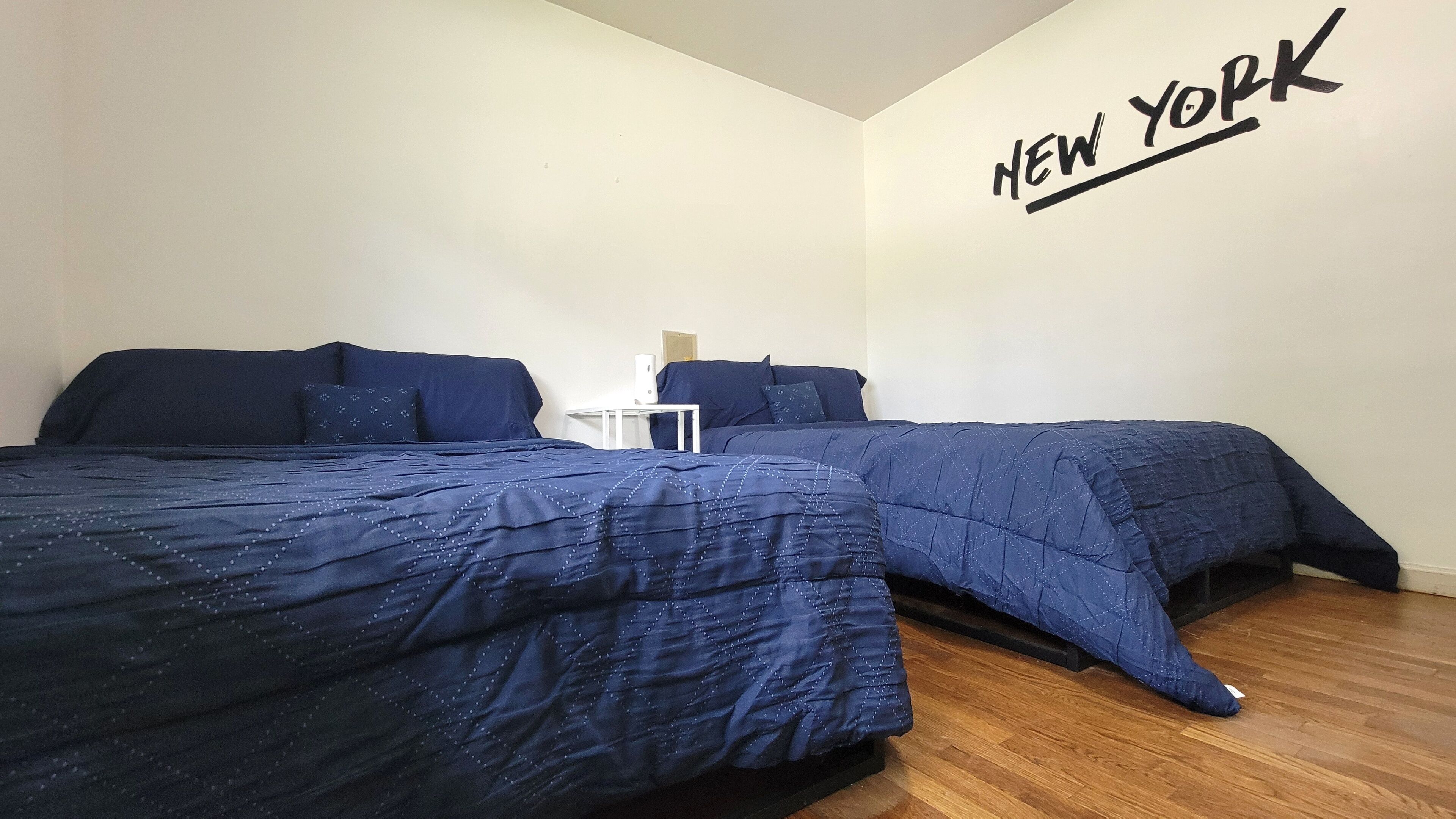 Une chambre avec deux lits, une table de chevet blanche et un mur sur lequel on peut lire le mot « NEW YORK ».