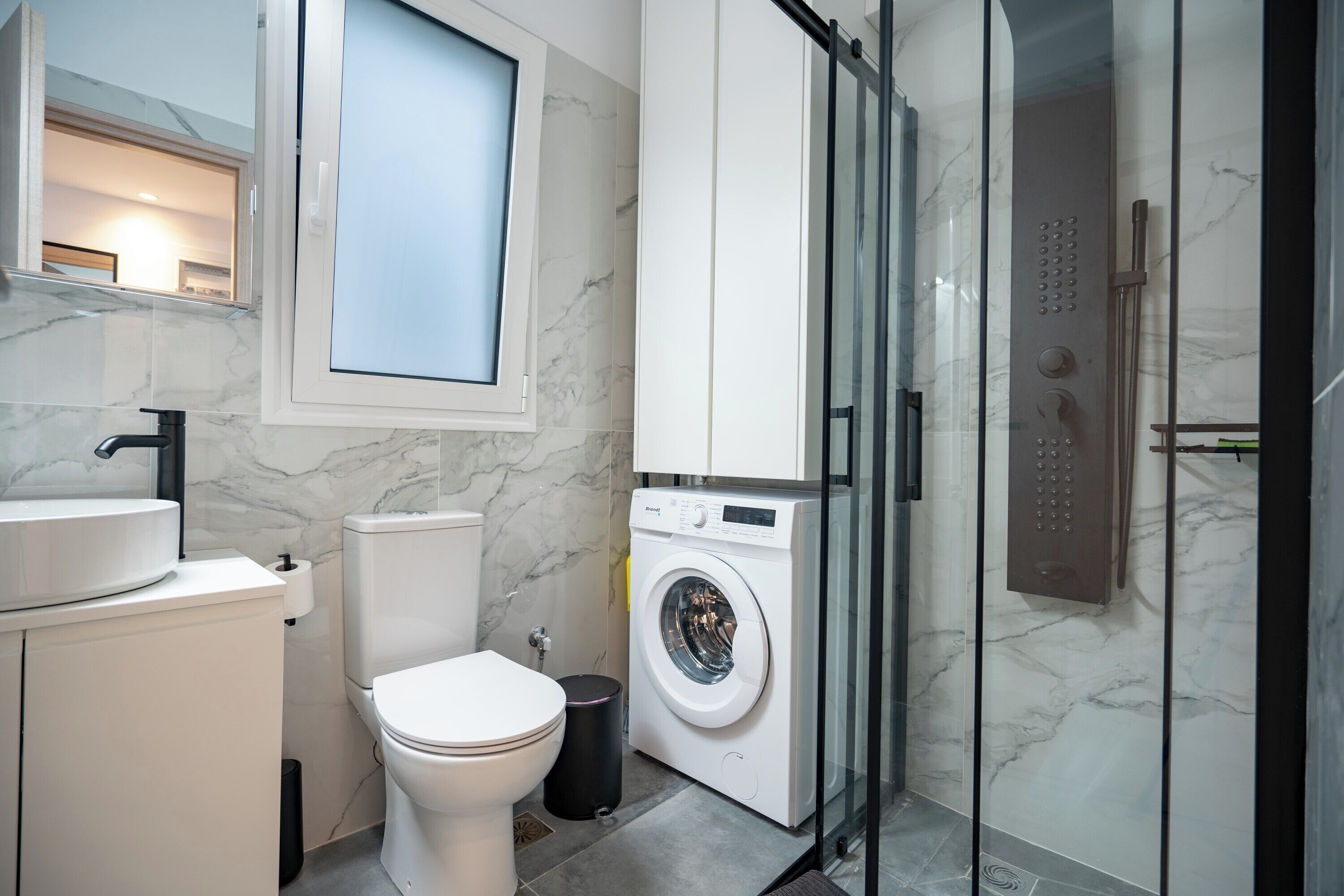 A modern bathroom with a toilet, washing machine, and glass-enclosed shower.