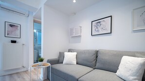 Ett modernt vardagsrum med en grå soffa, vita kuddar, ett litet runt bord med en växt och ett vitt element.