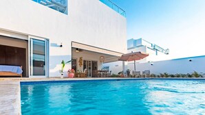 A modern poolside area with a clear blue pool, a covered seating area with umbrellas, and a large white building with a glass door and a staircase. - Villa Valentina Oceanfront Bliss (Sosúa)