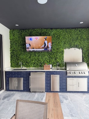 A modern outdoor kitchen with a flat-screen TV mounted on a green wall, a stainless steel grill, and a seating area with a woven chair.