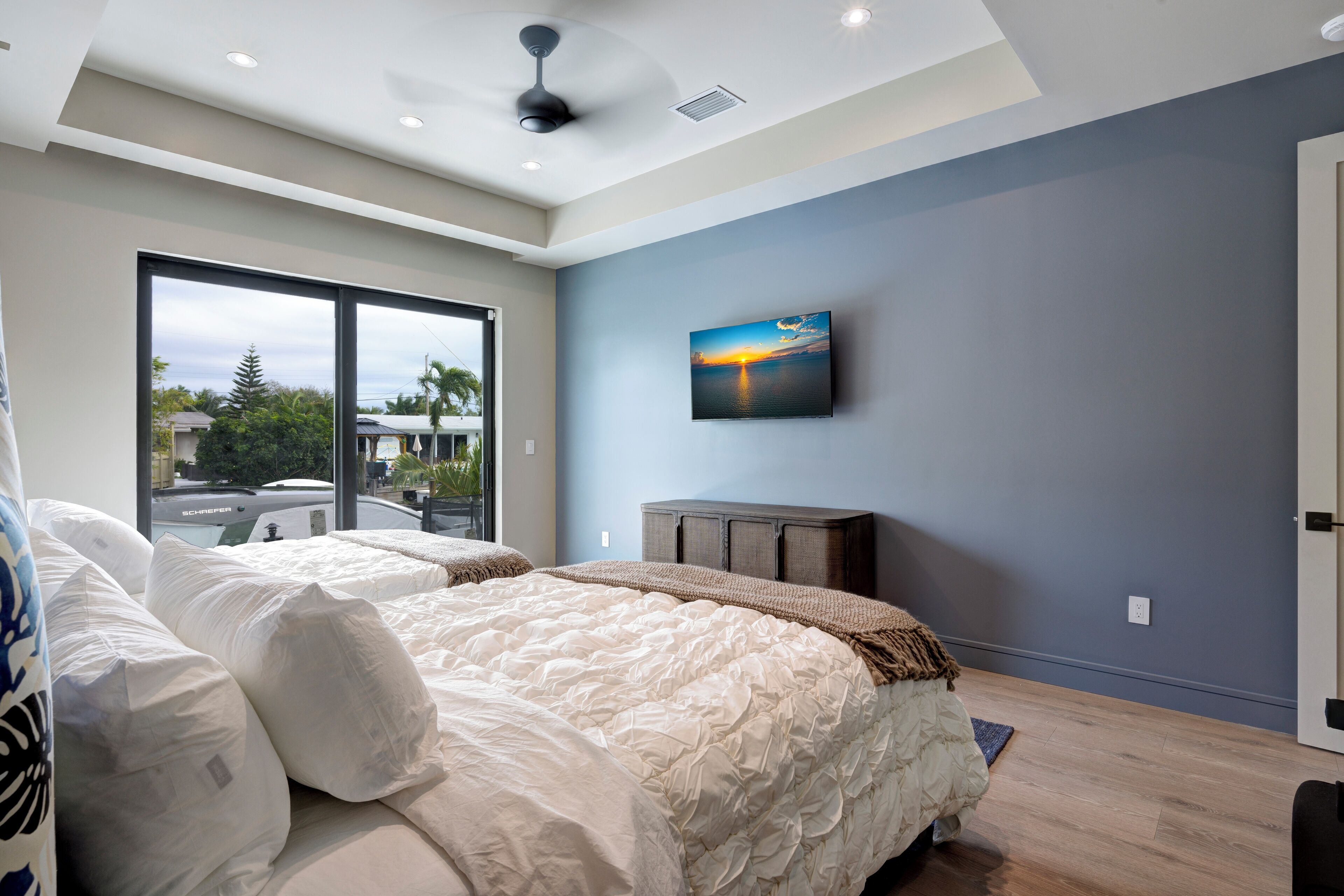 A bedroom with a large bed, a ceiling fan, a painting of a sunset, and a view of the outside.