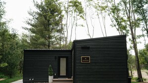 Una cabaña de madera negra con un pequeño porche y una puerta, rodeada de árboles y un camino de grava.